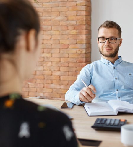 Business concept. Job interview. Adult bearded male hr manager in glasses and shirt sitting in light office in front of brunette caucasian woman, asking questions about previous work place.