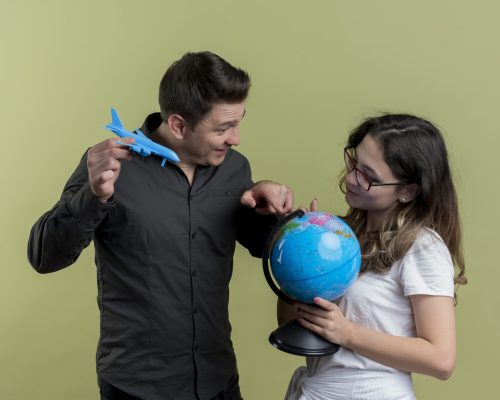 happy young couple of tourists man and woman holding globe and toy air plane standing together having fun over light background