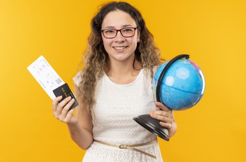 smiling young pretty schoolgirl wearing glasses holding airplane tickets, credit card and globe isolated on yellow background
